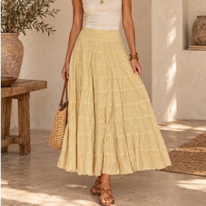 May include: A long, tiered skirt in a soft, pale yellow hue. The skirt features a high waistband and a full, flowing silhouette with horizontal seams creating multiple gathered layers that extend down to the hemline, providing a textured appearance.