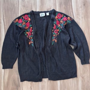 Puede incluir: Un cárdigan negro con bordado floral en rojo, naranja y verde. El cárdigan abierto tiene mangas largas y puños acanalados. El bordado floral está en los hombros y la parte superior del pecho. El cárdigan es de punto.