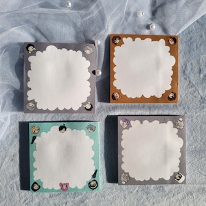 Peut inclure: Quatre blocs de notes adhésives carrés avec une zone d'écriture centrale blanche festonnée. Les blocs présentent des bordures grises, brunes et vert clair décorées de petites illustrations de personnages colorés aux coins et bords.