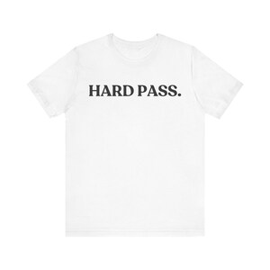 May include: A white short-sleeved t-shirt featuring the text "HARD PASS." printed in a bold, black, sans-serif font across the center of the chest. The garment has a classic crew neckline and a standard fit, showcasing a clean, minimalist design.