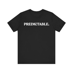 Può includere: T-shirt nero girocollo con la scritta "PREDICTABLE." stampata in bianco sul petto. La maglietta a maniche corte è realizzata in un materiale morbido.