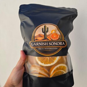 Puede incluir: Una bolsa negra de rodajas de naranja deshidratadas con una ventana transparente. La bolsa presenta un logotipo con una escena desértica y el texto "GARNISH SONORA" y "FRUTA DESHIDRATADA".