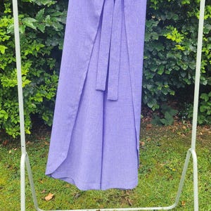 Puede incluir: Una falda envolvente de color lavanda con una cinta a juego, colgada en un perchero de metal blanco. La falda está hecha de una tela texturizada y tiene un diseño fluido. El fondo presenta follaje verde y hierba.