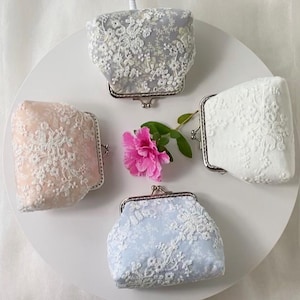 May include: Four small, lace-covered coin purses in pastel colors: light blue, pink, white, and gray. Each purse has a silver-colored clasp. A pink flower and green leaves are in the center of the purses, arranged on a white surface.