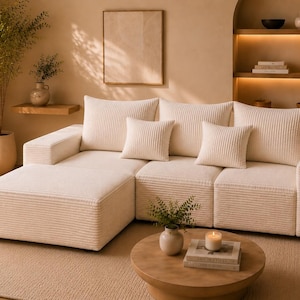 May include: A cream-colored sectional sofa with corduroy texture, featuring a chaise lounge and four square throw pillows. A round wooden coffee table holds a candle and books. The room has a neutral color palette.