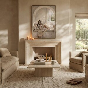 Person holding a framed neutral woman reading nook wall art print in a thin gold frame with no mat, showing the arched reading niche, draped linen, and warm beige tones.