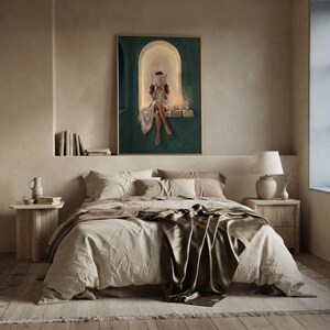 Emerald green reading nook art print hung above a bed made up in rumpled linen and olive silk, on a plaster bedroom wall beside a ledge of books and a ceramic lamp.