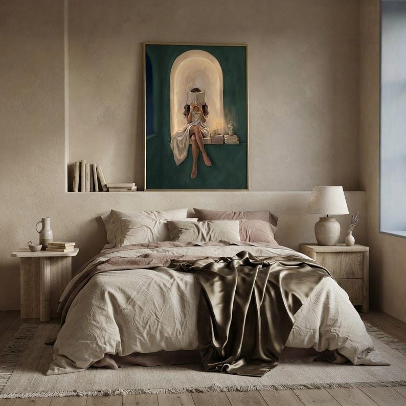 Emerald green reading nook art print hung above a bed made up in rumpled linen and olive silk, on a plaster bedroom wall beside a ledge of books and a ceramic lamp.