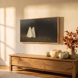 Two ghosts art from the set on a Frame TV in an autumn living room seen at an angle, with window-pane sunlight, an oak bench with pumpkins and a knit throw
