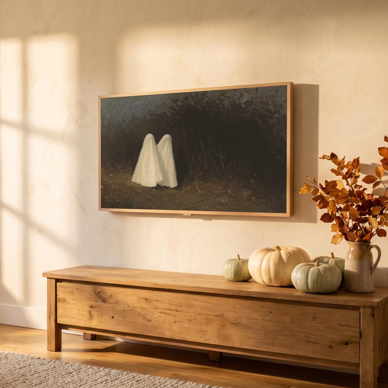 Two ghosts art from the set on a Frame TV in an autumn living room seen at an angle, with window-pane sunlight, an oak bench with pumpkins and a knit throw