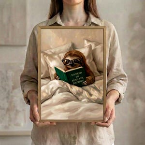 Person in a linen shirt holding the framed sloth reading print in a slim gold frame, showing its true scale against a soft neutral background