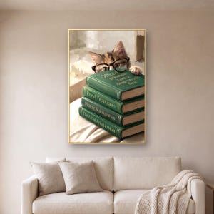 The cat peeking over books print in a thin gold frame on a warm plaster wall above a cream linen sofa with two cushions and a knitted throw over one arm, in soft daylight.