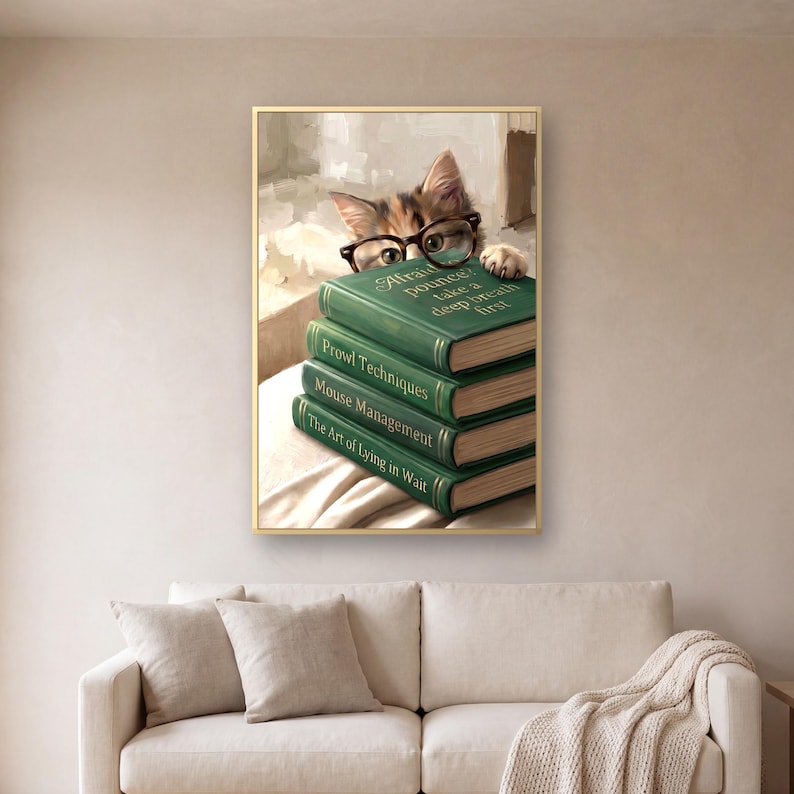 The cat peeking over books print in a thin gold frame on a warm plaster wall above a cream linen sofa with two cushions and a knitted throw over one arm, in soft daylight.