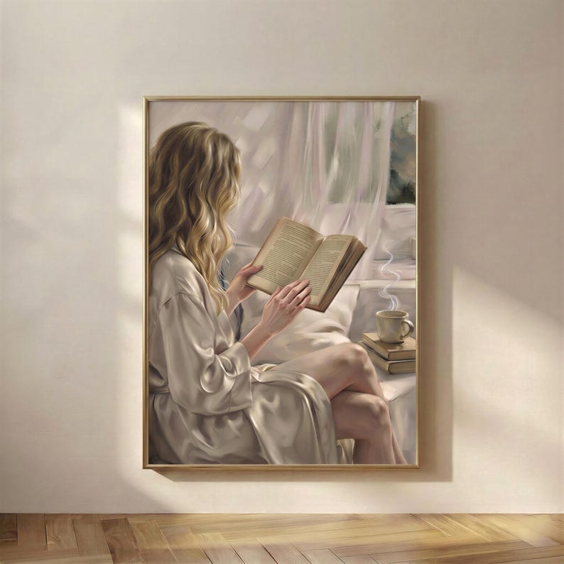Framed art print of a blonde woman reading a book in soft daylight, hanging in a thin gold frame on a warm cream plaster wall above a wooden floor