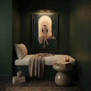 Emerald green reading nook art print lit by a brass picture light in a dark green alcove, above a cushioned bench with a knit throw, a candle, a mug and stacked books.