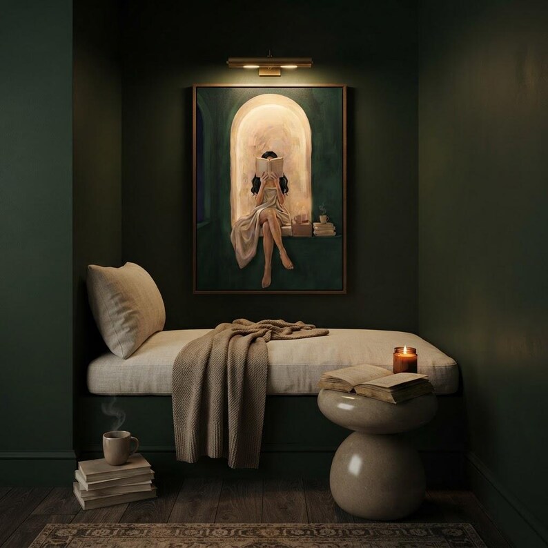 Emerald green reading nook art print lit by a brass picture light in a dark green alcove, above a cushioned bench with a knit throw, a candle, a mug and stacked books.