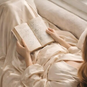 Close-up of the Quiet Reader printable wall art: a woman in a satin robe reading a book in a window seat nook, viewed from directly above, warm beige tones.