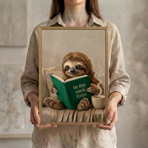A person in a linen shirt holding the sloth reading art print in a slim gold frame against a soft neutral background
