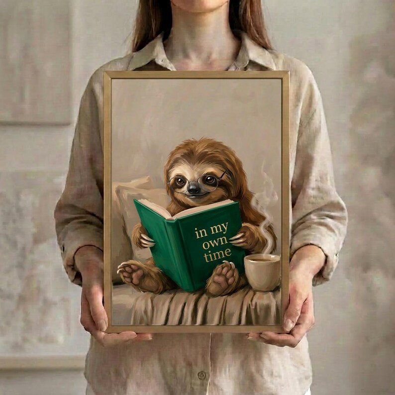 A person in a linen shirt holding the sloth reading art print in a slim gold frame against a soft neutral background