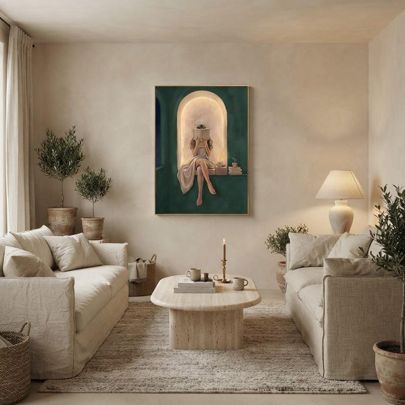 Emerald green reading nook art print in a thin gold frame on a warm plaster wall, between two cream linen sofas with a travertine coffee table, potted olive trees and a lit taper candle.