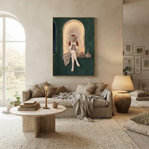 Emerald green reading nook art print in a thin gold frame on a plaster wall above a taupe linen sofa, with a round travertine table, a lit taper, stacked books and a tall arched garden window.