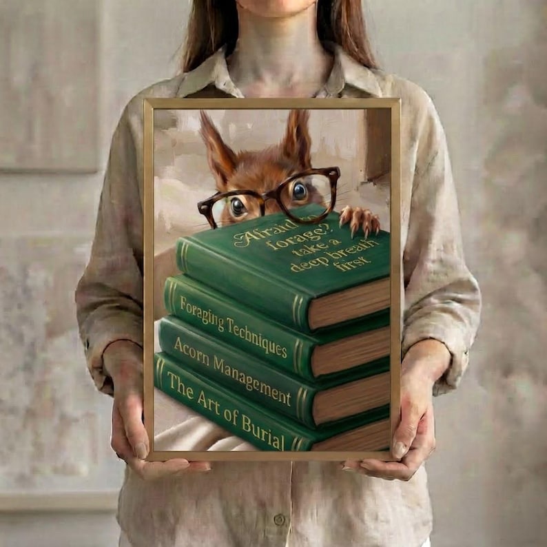 A person in a linen shirt holding the squirrel peeking over books art print in a slim gold frame against a soft neutral background