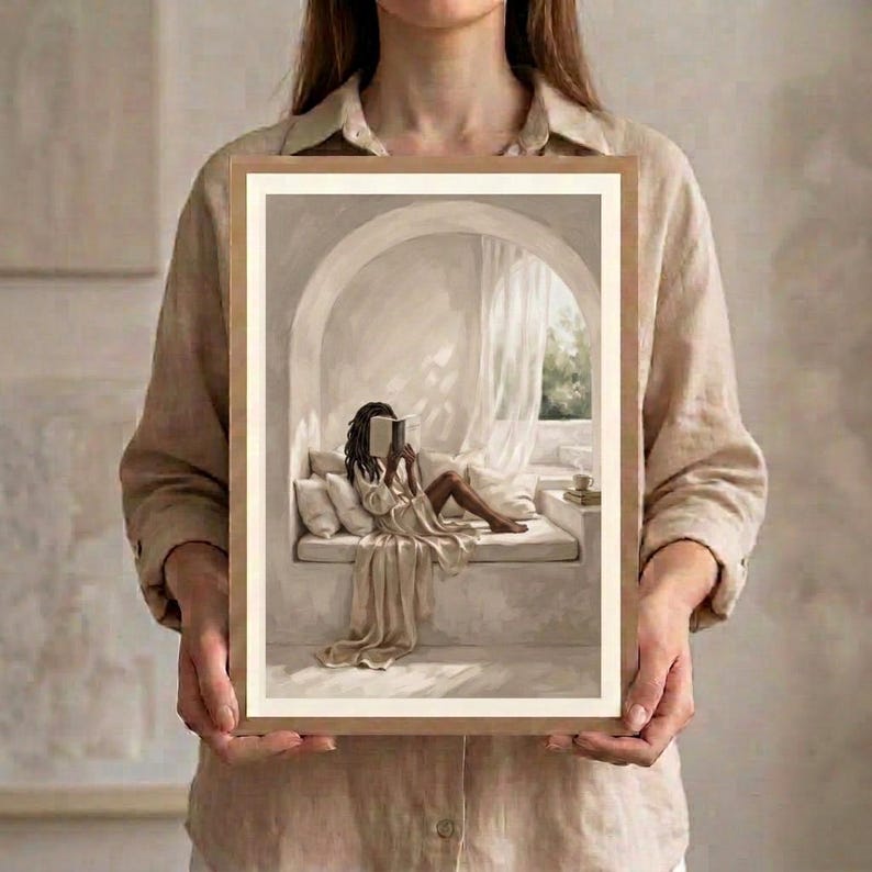 Hands holding a framed neutral Black woman reading nook wall art print in a thin gold frame with no mat against a light wall, showing the arched reading niche, draped linen, and warm beige tones.