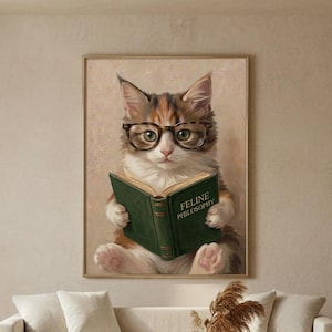 Painterly print of a calico kitten in round tortoiseshell glasses reading a green book framed in a slim wood frame on a warm plaster living-room wall above a cream linen sofa