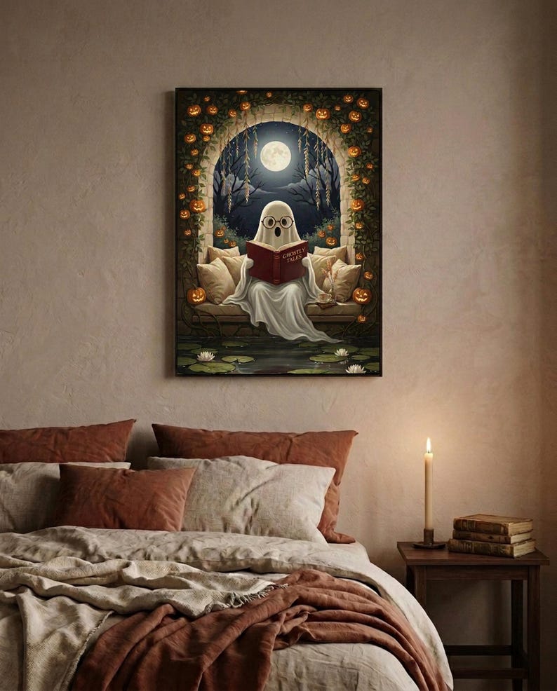 The pumpkin arch ghost reading print framed above a bed with linen bedding and a lit candle on the nightstand