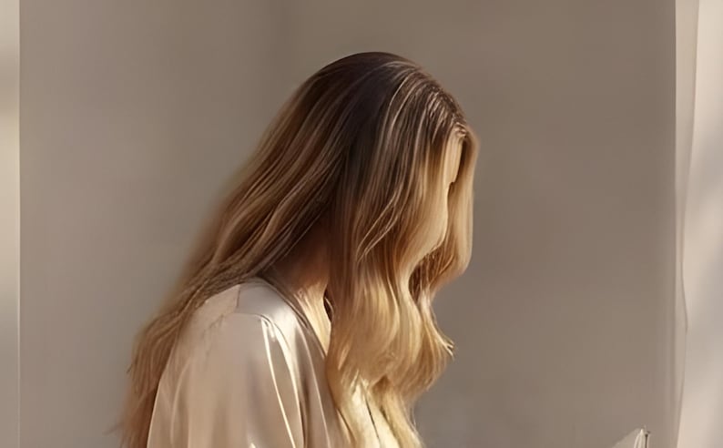 Close-up detail from Woman Reading Wall Art, a cropped fragment of the printable artwork: long blonde hair falling in loose waves over a satin robe, sunlight catching the strands.