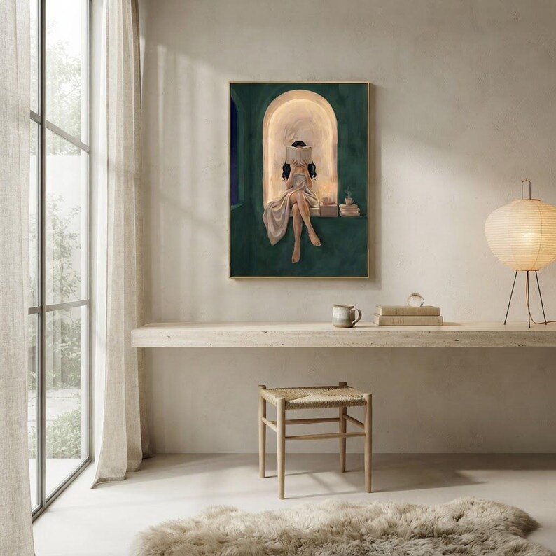 Emerald green reading nook art print on a pale plaster wall above a floating travertine console with a mug and books, beside a paper lantern lamp and a tall garden window.