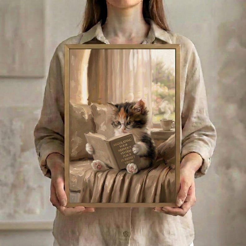 A person in a linen shirt holding the cat reading art print in a slim gold frame against a soft neutral background
