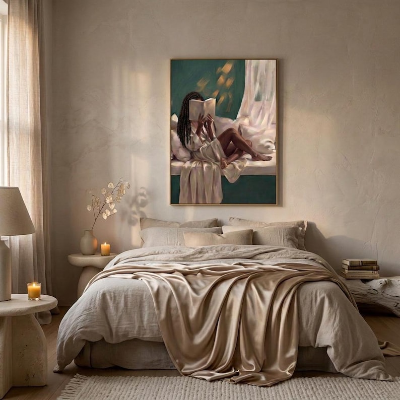 Gold-framed emerald green art print of a Black woman reading on a window seat above a bed layered in soft linen, a bedside lamp and candle glowing in a calm neutral bedroom.