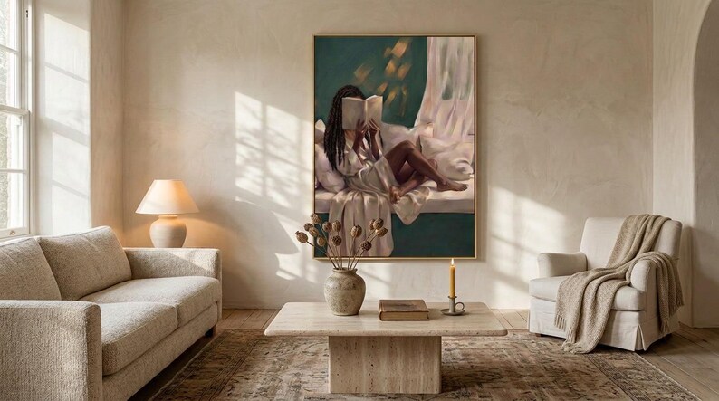 Gold-framed emerald green art print of a Black woman reading on a window seat in a sunlit living room with a linen sofa and armchair, shown wide to give a sense of scale.