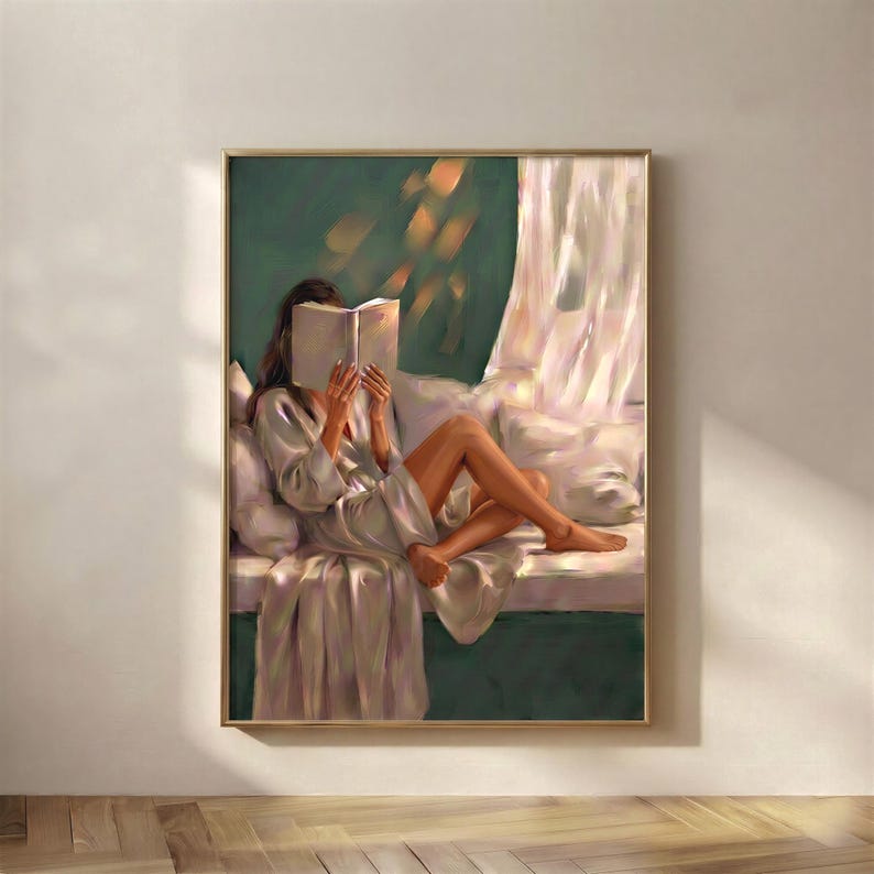 Emerald green wall art print in a thin gold frame on a warm white plaster wall above a herringbone wood floor, showing a brunette woman in a satin robe reading a book on a window seat with a sheer curtain and soft amber light.