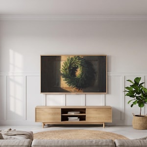 May include: A rectangular television screen displaying a digital painting of a lush green pine wreath hanging on a dark wooden door, mounted on a white paneled wall above a light oak media console with two closed cabinets and open shelving.