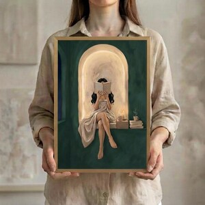The woman with long straight dark hair reading in an emerald green arched nook art print in a thin gold frame, held up in both hands to show its size.