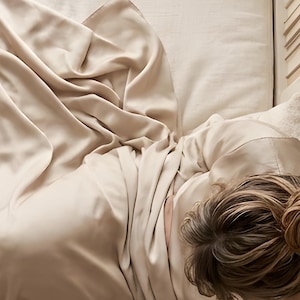 Close-up detail from Reading in Bed Wall Art, a cropped fragment of the printable artwork: rumpled silk and linen bedding and a loose low bun, seen from above.