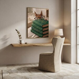Squirrel in round glasses peeking over books print framed above a restrained workspace with pale walls and a natural linen chair