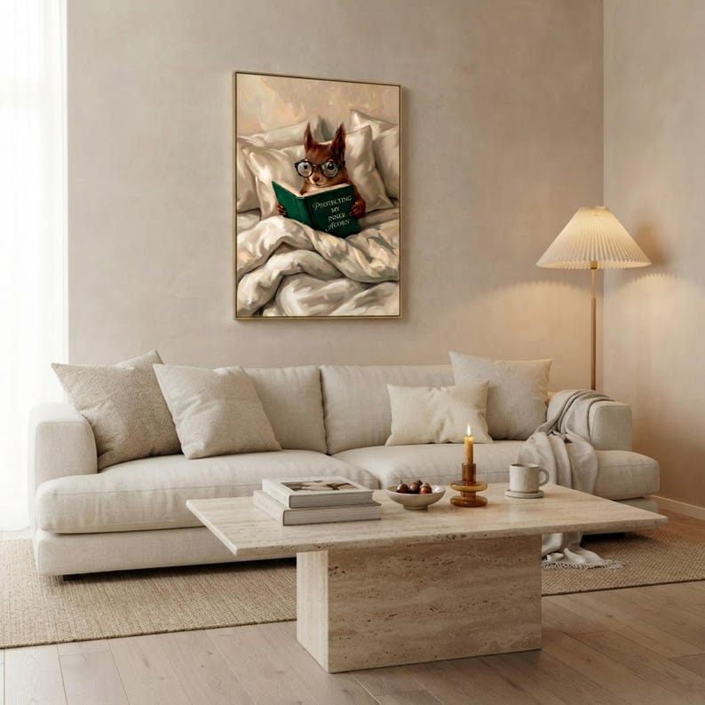 The squirrel reading print framed above a long cream sofa in a calm living room with a travertine coffee table and a floor lamp with a pleated shade