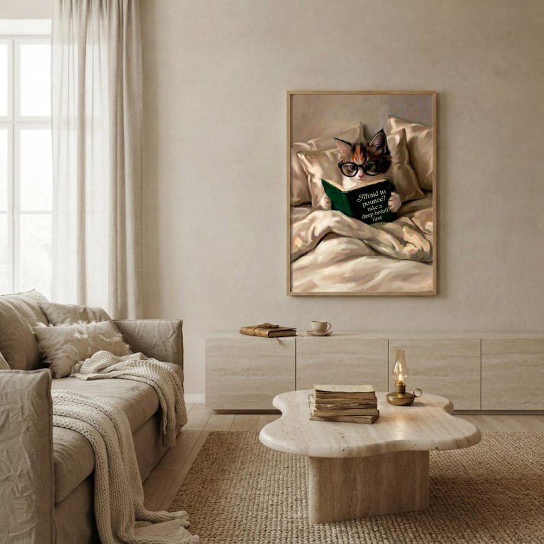 The framed cat reading print hanging above a cream sofa in a calm, light-filled living room