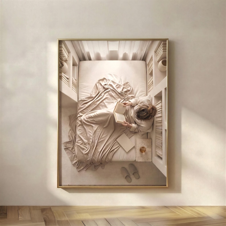Framed print of an overhead view of a linen-dressed bed, a figure reading under soft sheets with books either side, hung on a warm plaster wall above a pale oak herringbone floor. neutral printable wall art, minimalist home decor, Japandi style, calm living room and bedroom decor, slow living digital download.