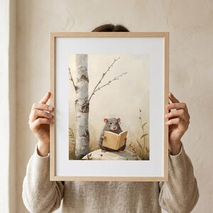 A person in an oatmeal linen sweater holding up a framed woodland nursery print of a mouse in round glasses reading a book, shown at arm's length against a warm cream wall