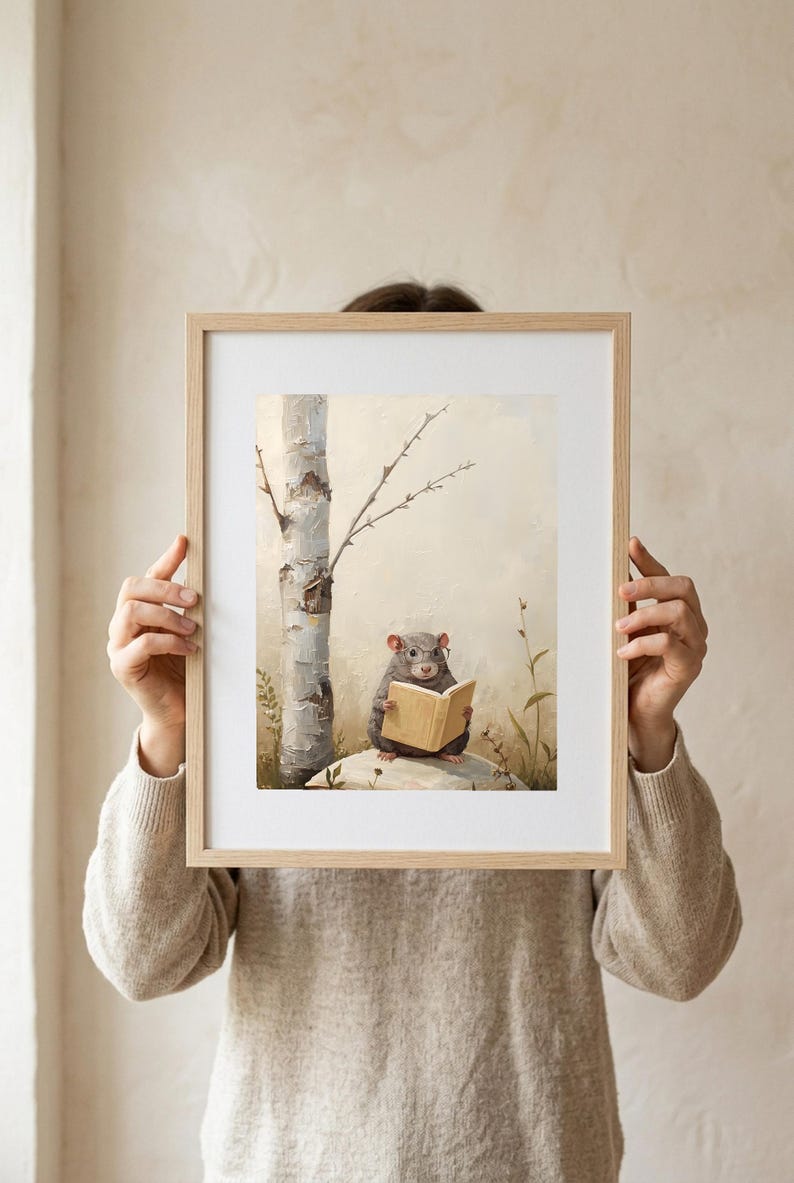 A person in an oatmeal linen sweater holding up a framed woodland nursery print of a mouse in round glasses reading a book, shown at arm's length against a warm cream wall