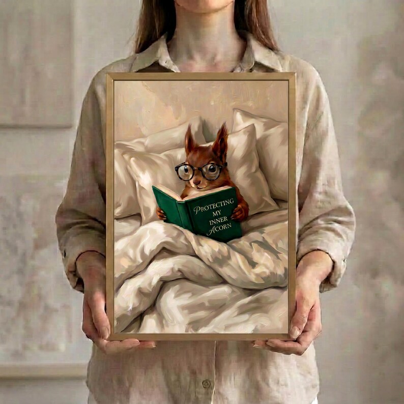Person in a linen shirt holding the framed squirrel reading print in a slim gold frame, showing its true scale against a soft neutral background