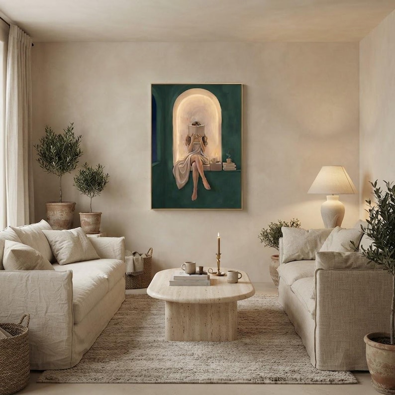 Emerald green reading nook art print in a thin gold frame on a warm plaster wall, between two cream linen sofas with a travertine coffee table, potted olive trees and a lit taper candle.