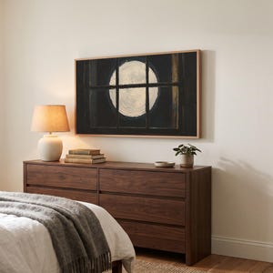 Moon through window panes art from the set on a Frame TV in a bedroom seen at an angle, above a walnut dresser with a lit lamp, beside a linen bed and throw