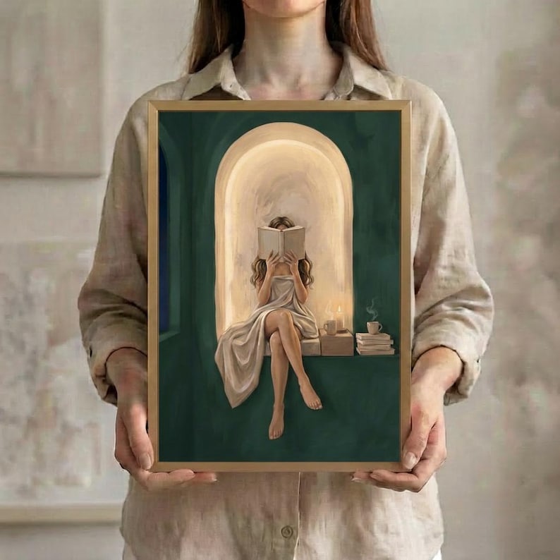 The emerald green arched nook art print of the woman with light brown hair reading, in a thin gold frame, held up in both hands to show its size.