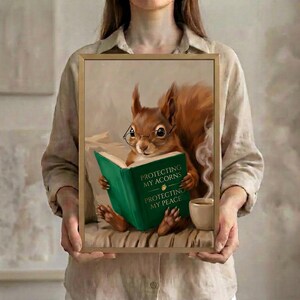 A person in a linen shirt holding the red squirrel reading art print in a slim gold frame against a soft neutral background