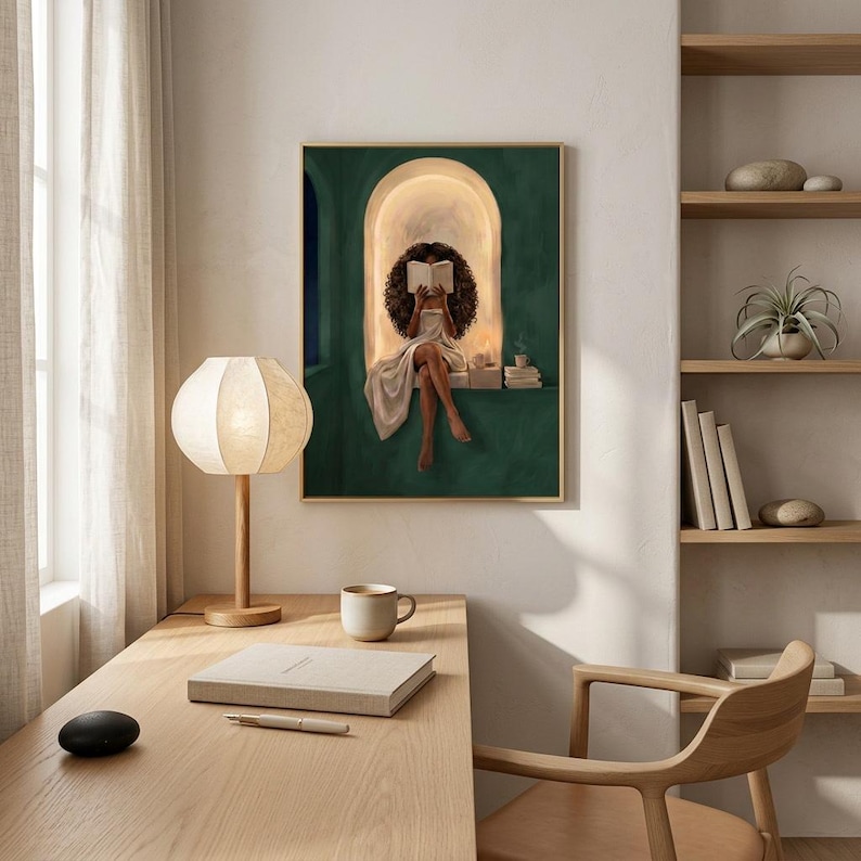 Emerald green reading nook art print in a thin gold frame above a light oak desk with a paper lantern lamp, a coffee cup and a linen notebook, beside open shelving in a sunlit study.